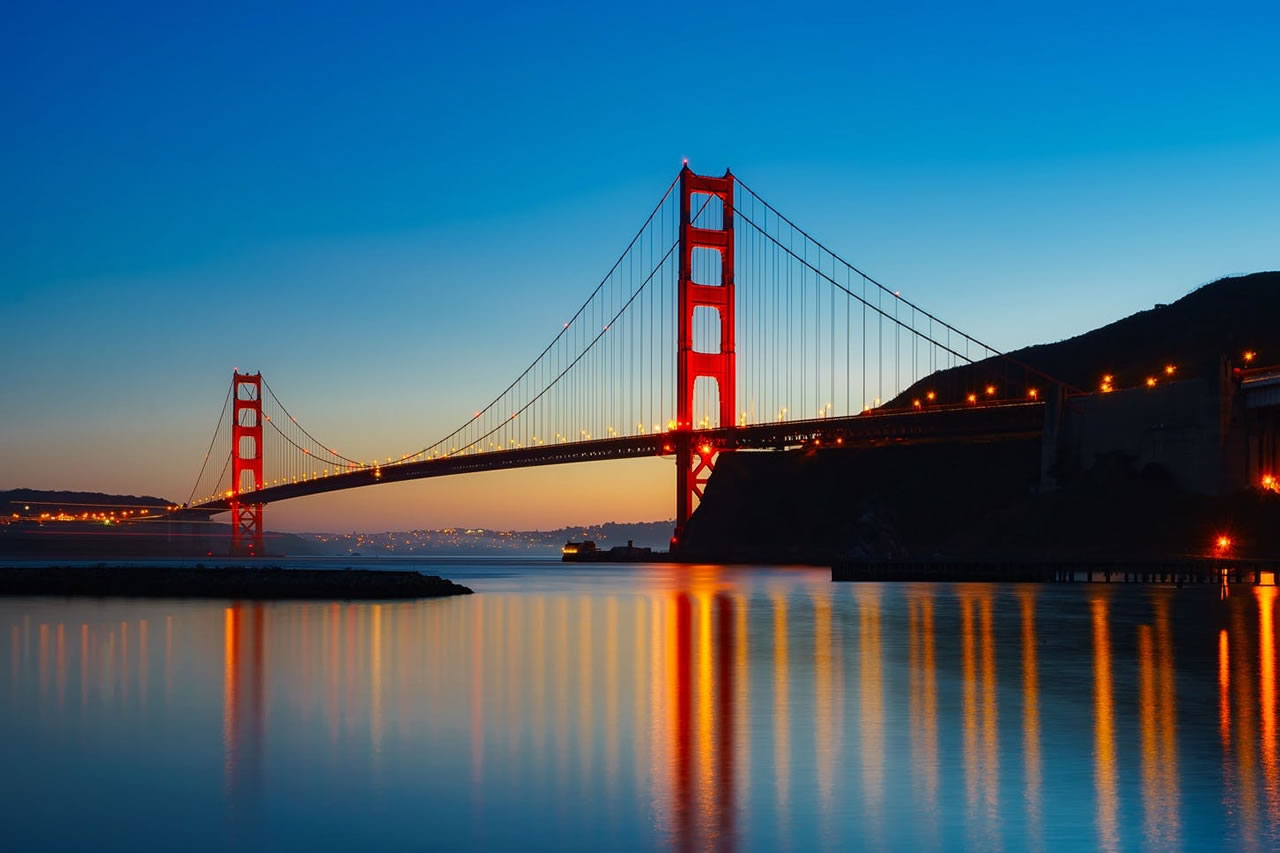 Image of the Golden Gate Bridge at sunset. The sky is a deep blue, and the bridge is lit up with orange lights.