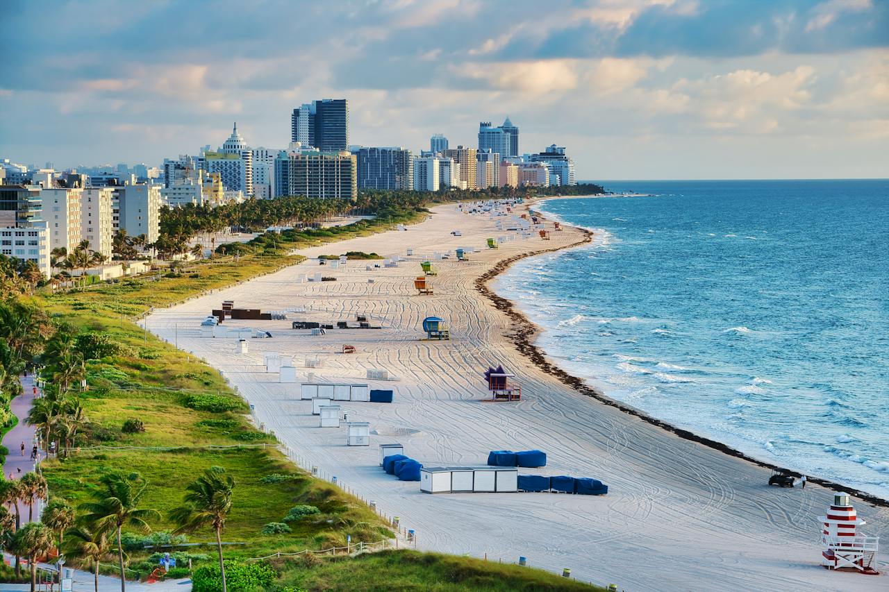 Image of a beach with clear blue water and white sand. There are palm trees in the background.